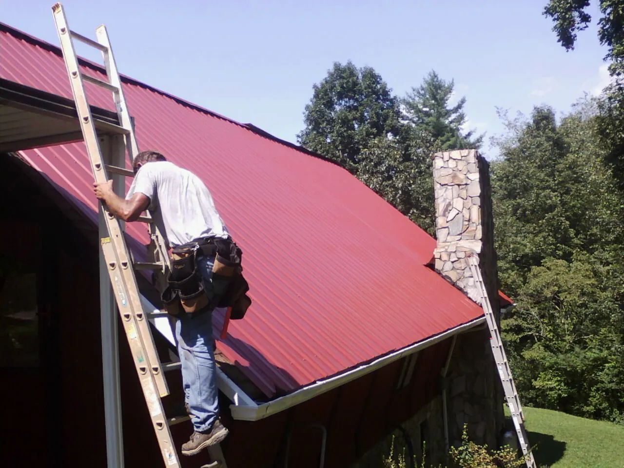 Roofer on a ladder beside a red metal roof and stone chimney — Soffit & Fascia Repair by Roofing Brothers in Craigsville
